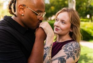 A man kisses a tattooed woman's hand outdoors in a romantic gesture, with green trees in the background.