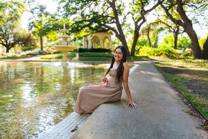 A young woman in a flowing beige dress sits gracefully by a serene garden pond with a fountain and lush trees.