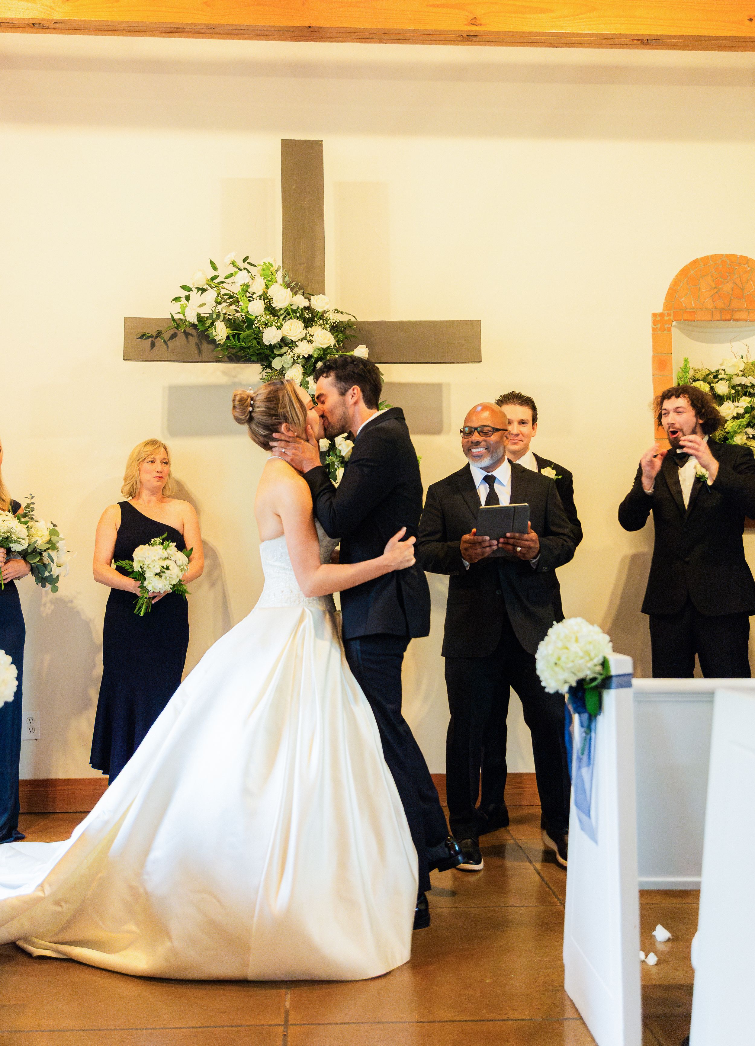 Bride and groom share first kiss at wedding altar with cross, flowers, and wedding party in church ceremony.