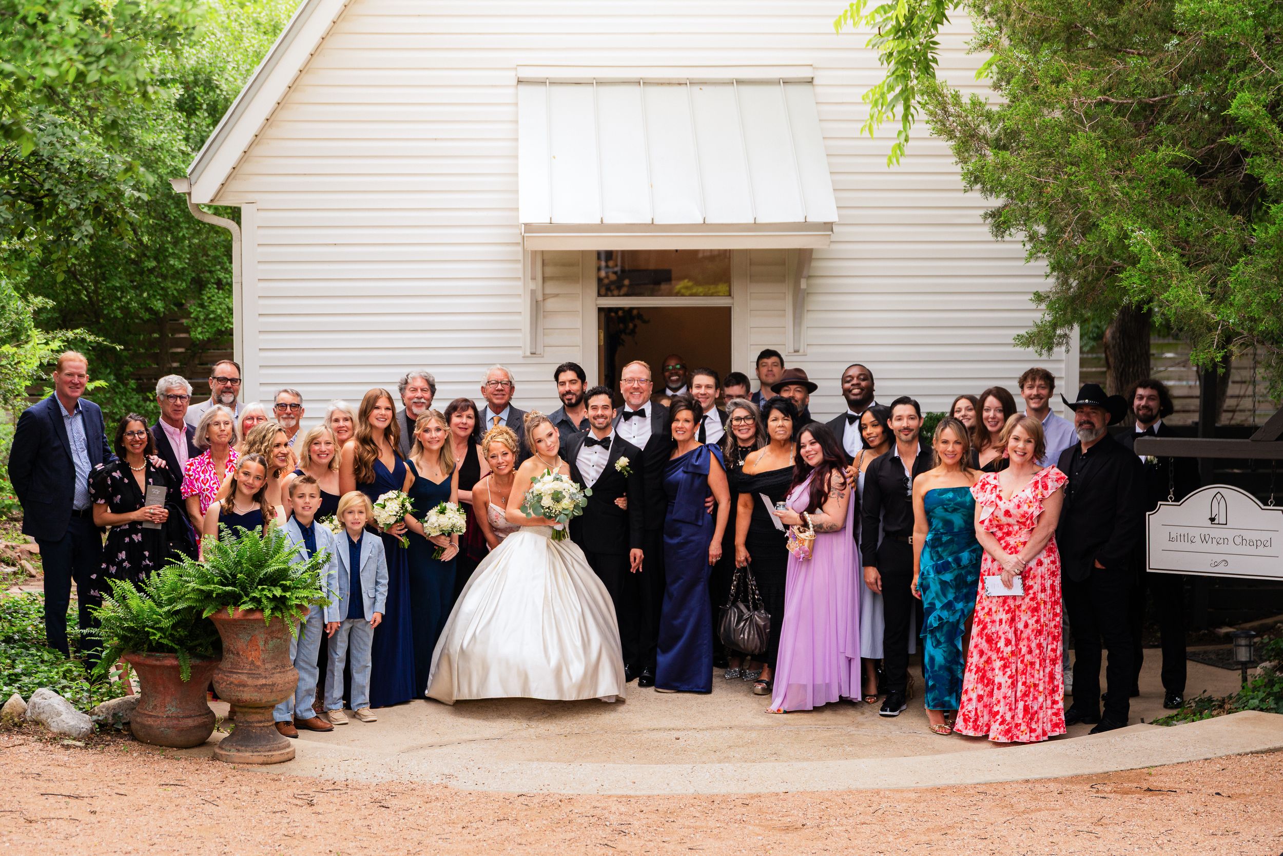 Wedding party and guests pose for a group photo outside a charming white chapel venue.