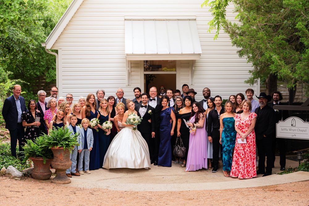 Wedding party and guests pose for a group photo outside a charming white chapel venue.