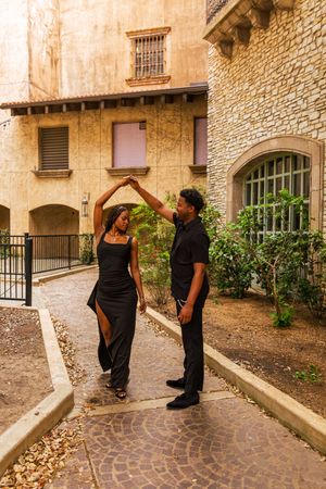 A couple in black outfits dancing outdoors, man spinning woman in elegant dress near stone buildings.