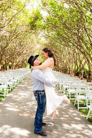 Groom in cowboy hat lifts bride in white dress outdoors at tree-lined wedding ceremony aisle with white chairs.