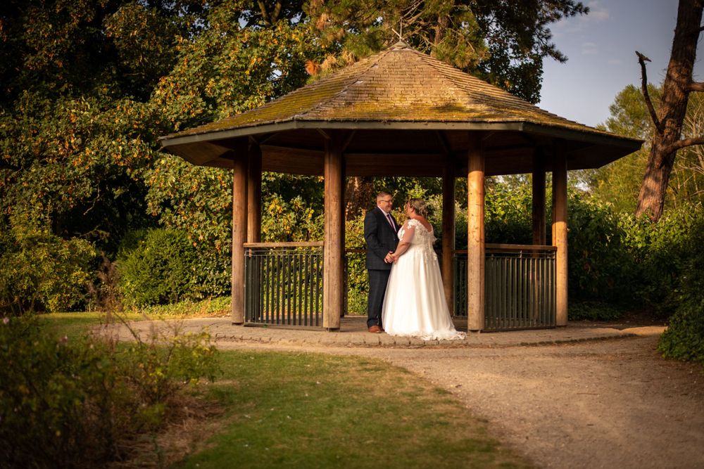 Elegant wedding couple standing in a wooden gazebo surrounded by lush greenery at sunset.