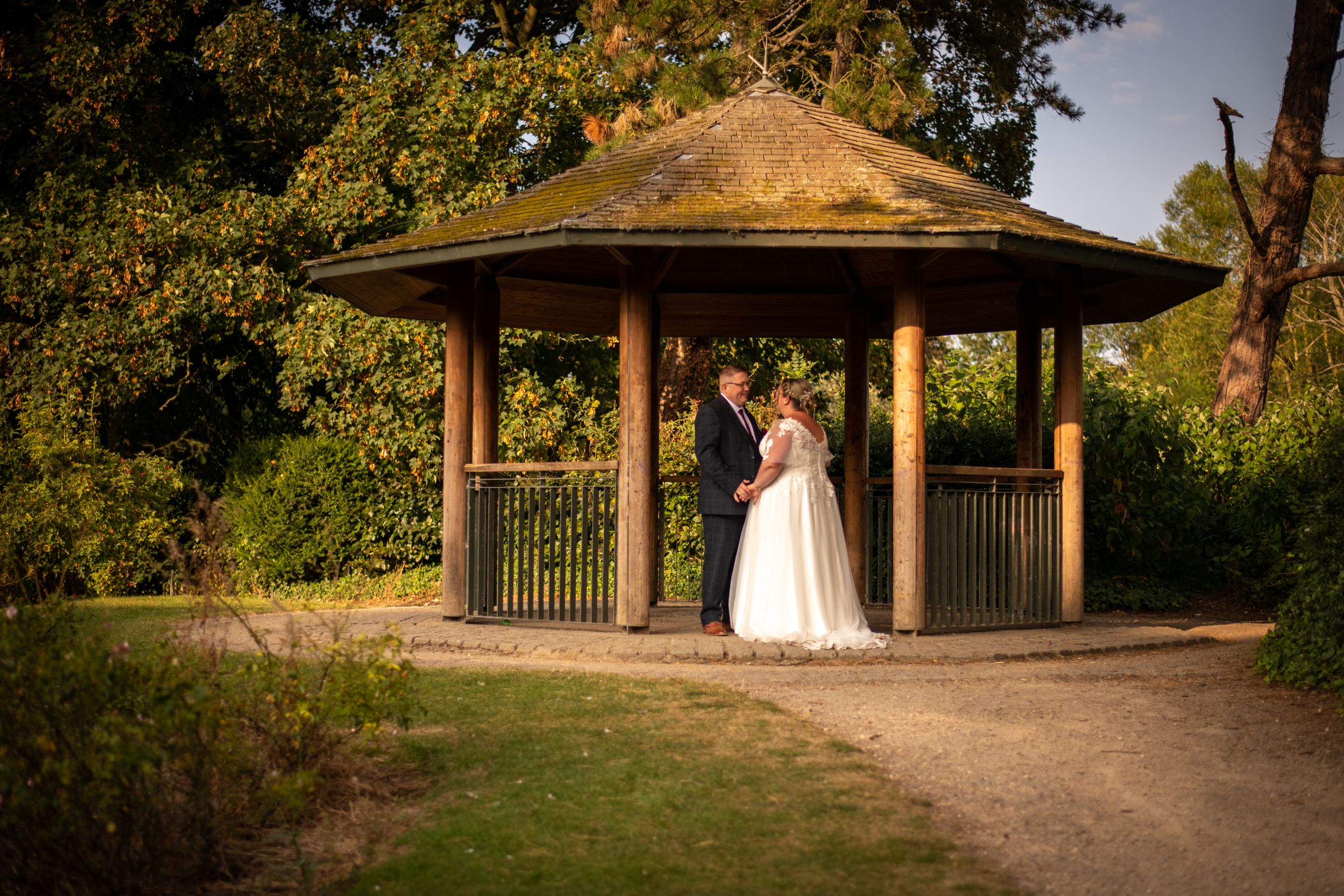 Elegant wedding couple standing in a wooden gazebo surrounded by lush greenery at sunset.