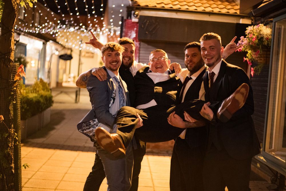 Wedding party lifts someone up while celebrating under outdoor string lights at dusk.