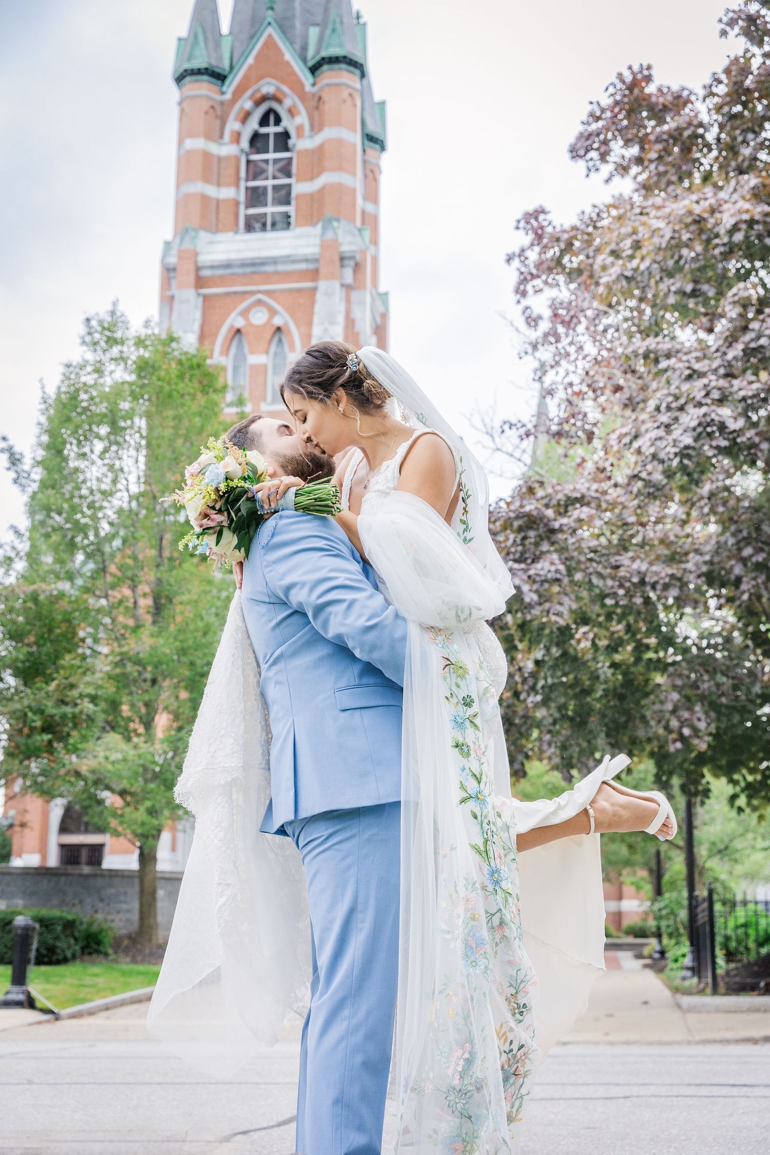 A romantic wedding moment captured in front of a Gothic church tower with blooming spring trees.