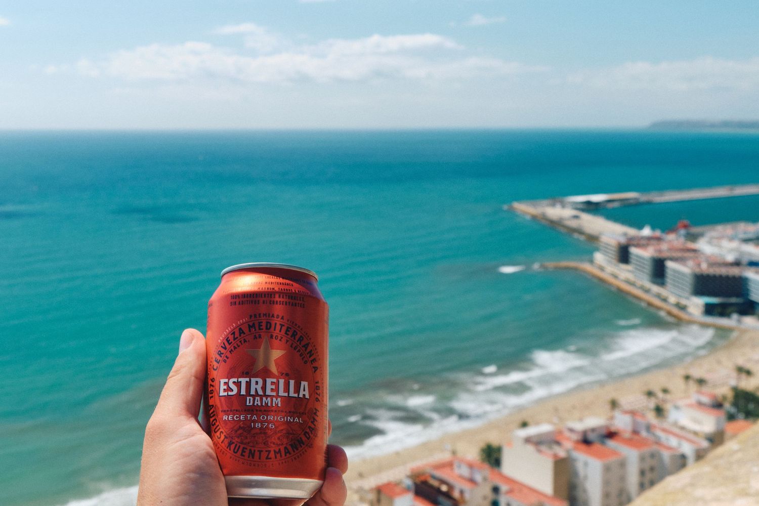 Red Estrella beer can held up against a scenic coastal Mediterranean beach view.