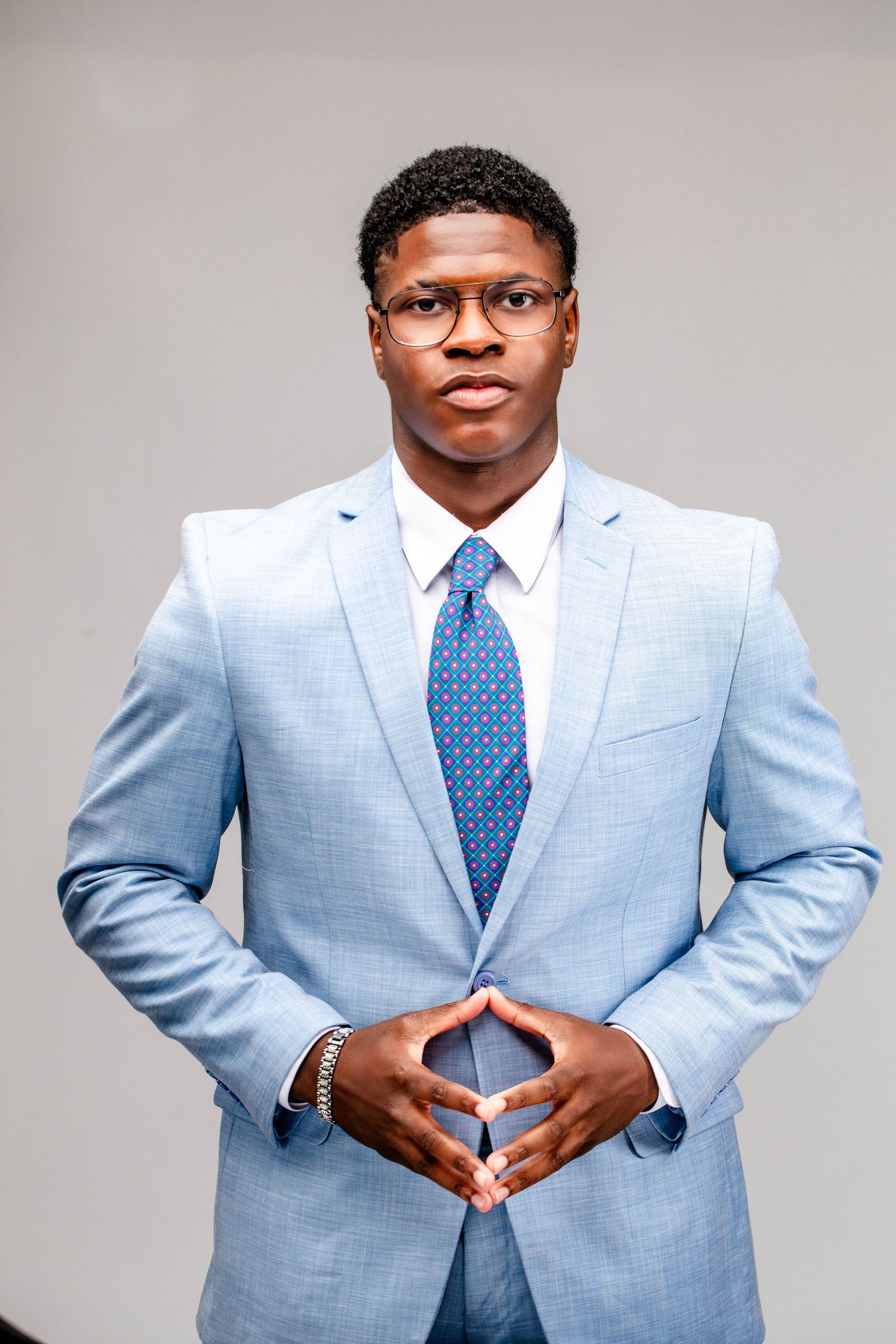 Young man in round glasses and a light blue suit with a patterned tie poses with fingertips pressed together against a gray background.