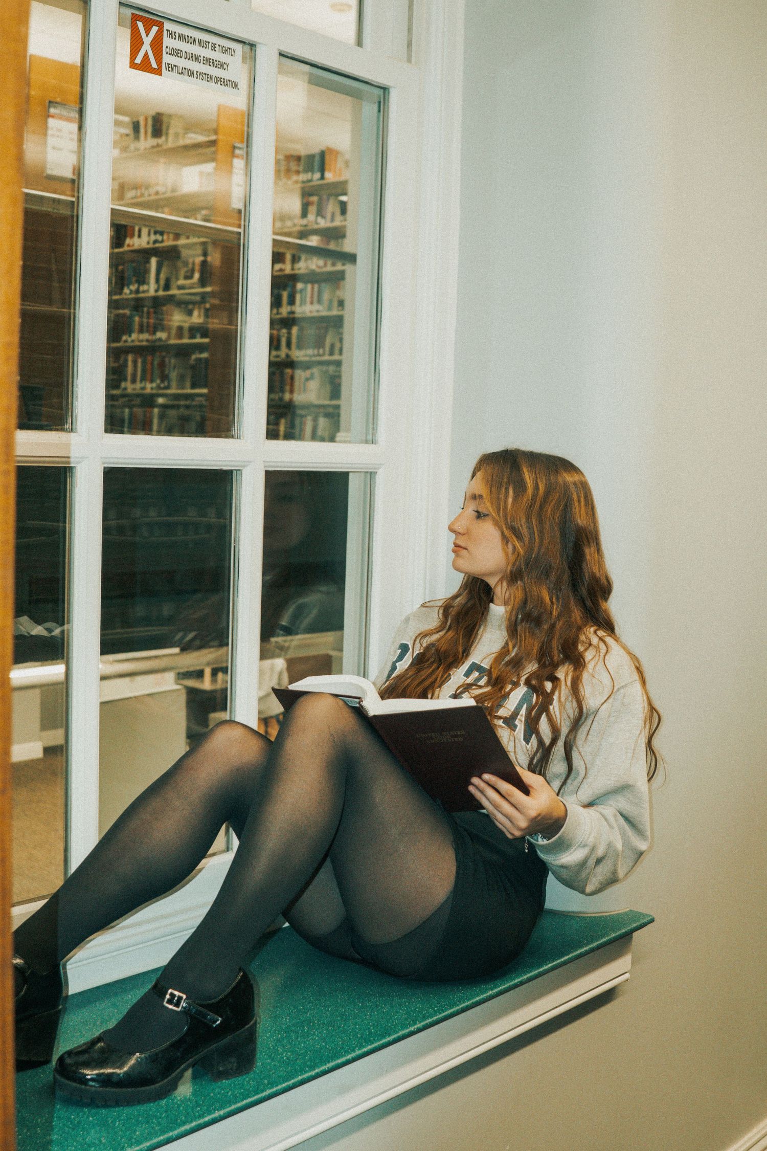 Student sitting by library window ledge in white sweater and black tights while reading a book.