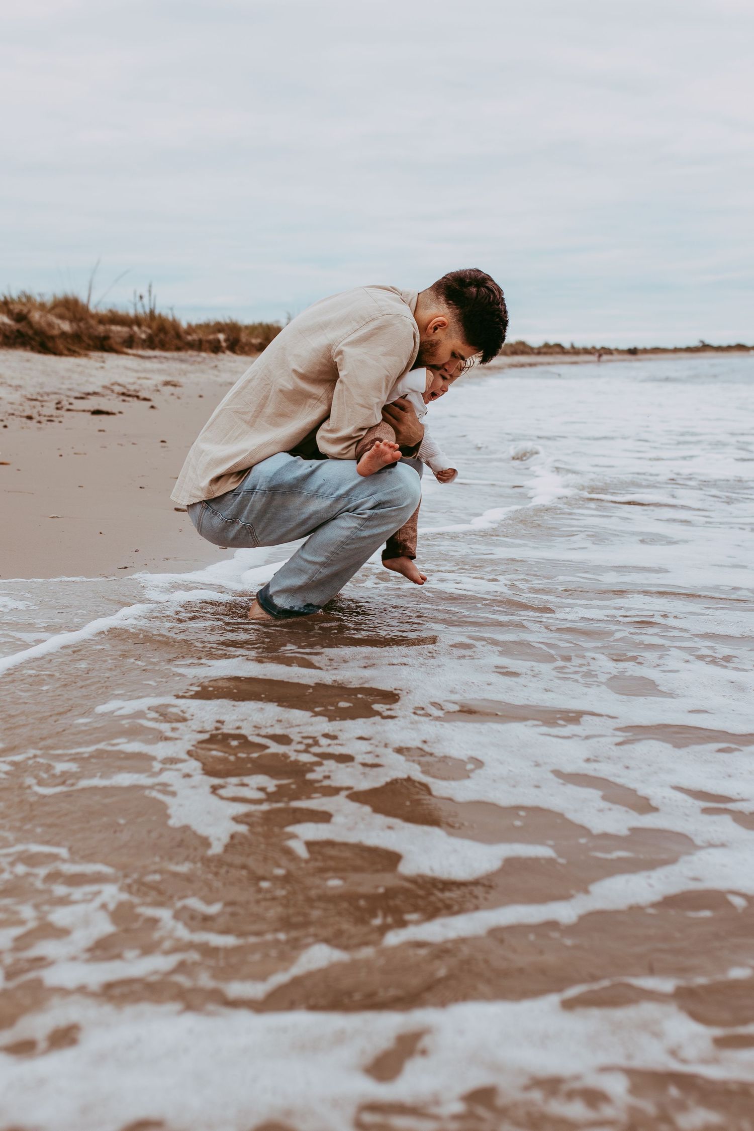 Father crouching at the edge of the ocean shore as waves wash up onto the sandy beach during an overcast day. Whitney Popp, photographer