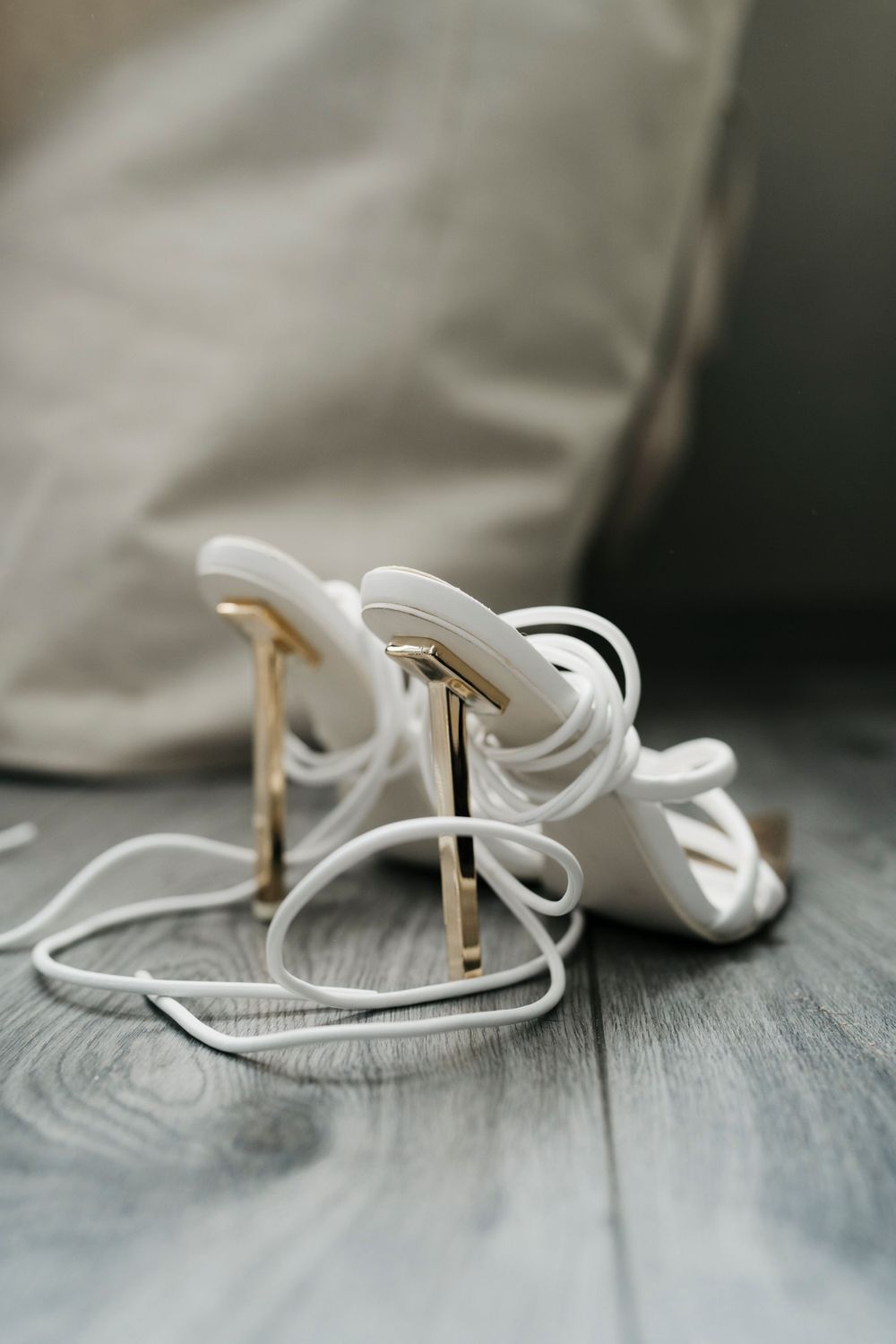 White strappy high heel sandals with gold stiletto heels on wooden floor.
