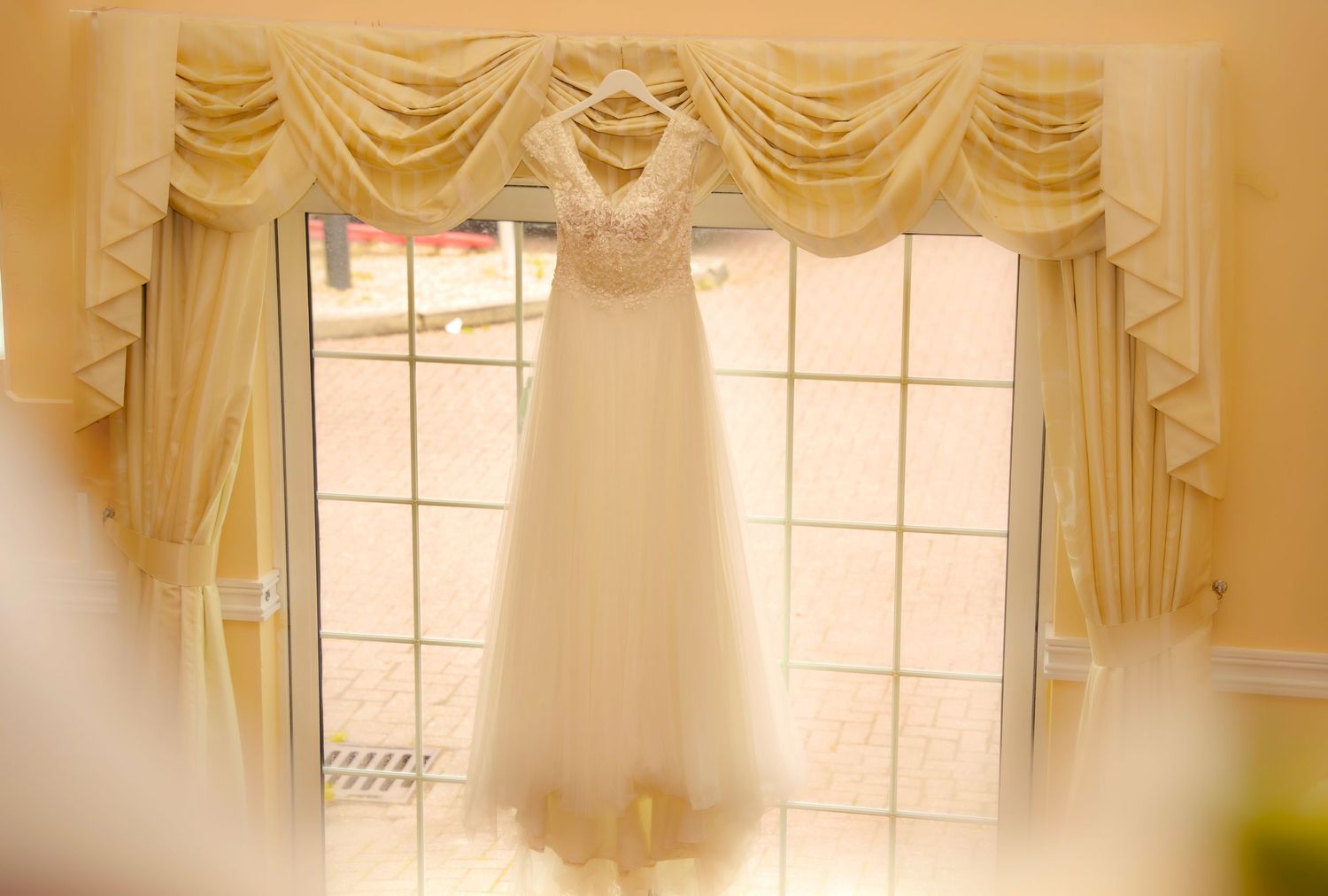 Elegant cream colored wedding dress hanging by window with golden curtains and natural light.