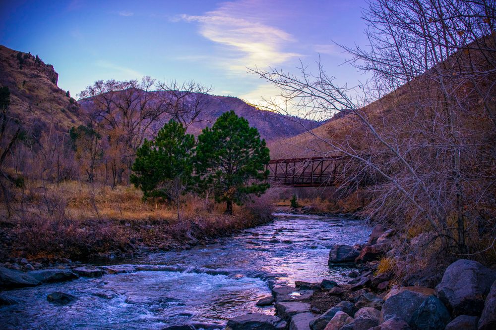 Rustic bridge spans a rushing mountain stream at sunset with rocky cliffs.