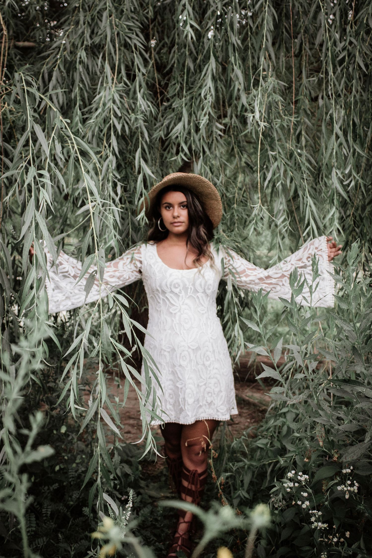 A figure in a white lace dress and brown boots poses among drooping willow branches.