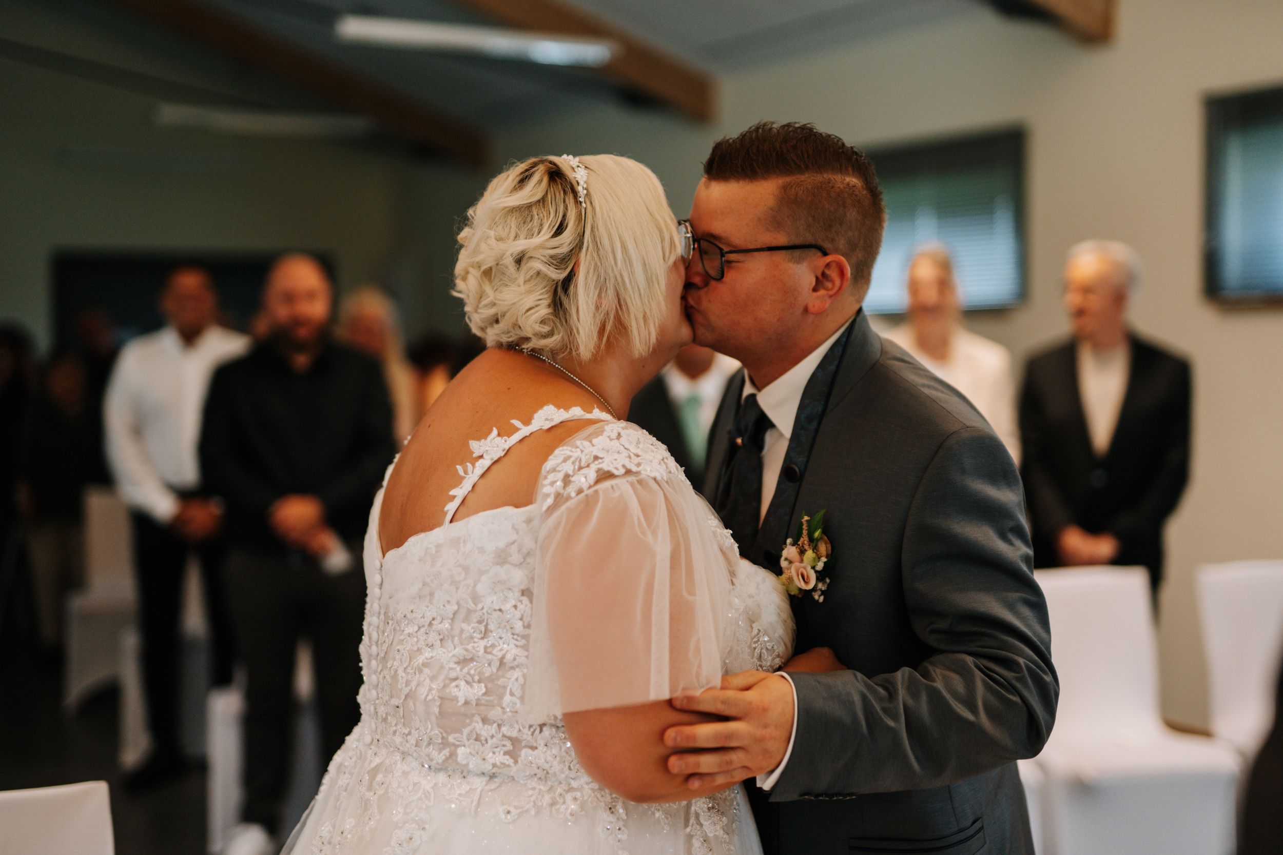 Intimate moment during indoor wedding ceremony with guests in background.
