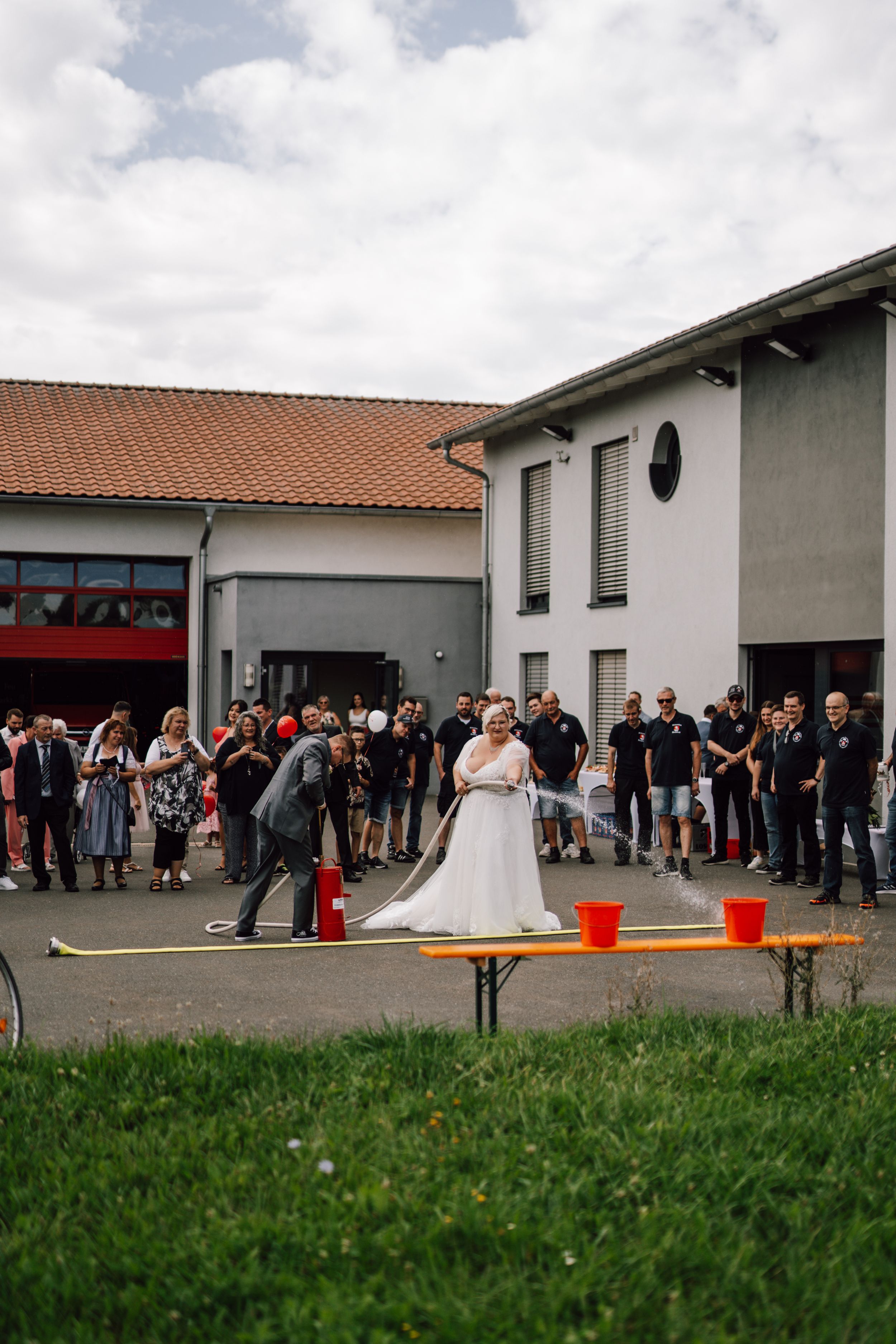 Wedding guests gather outside a building with red decorations on a grassy lawn.