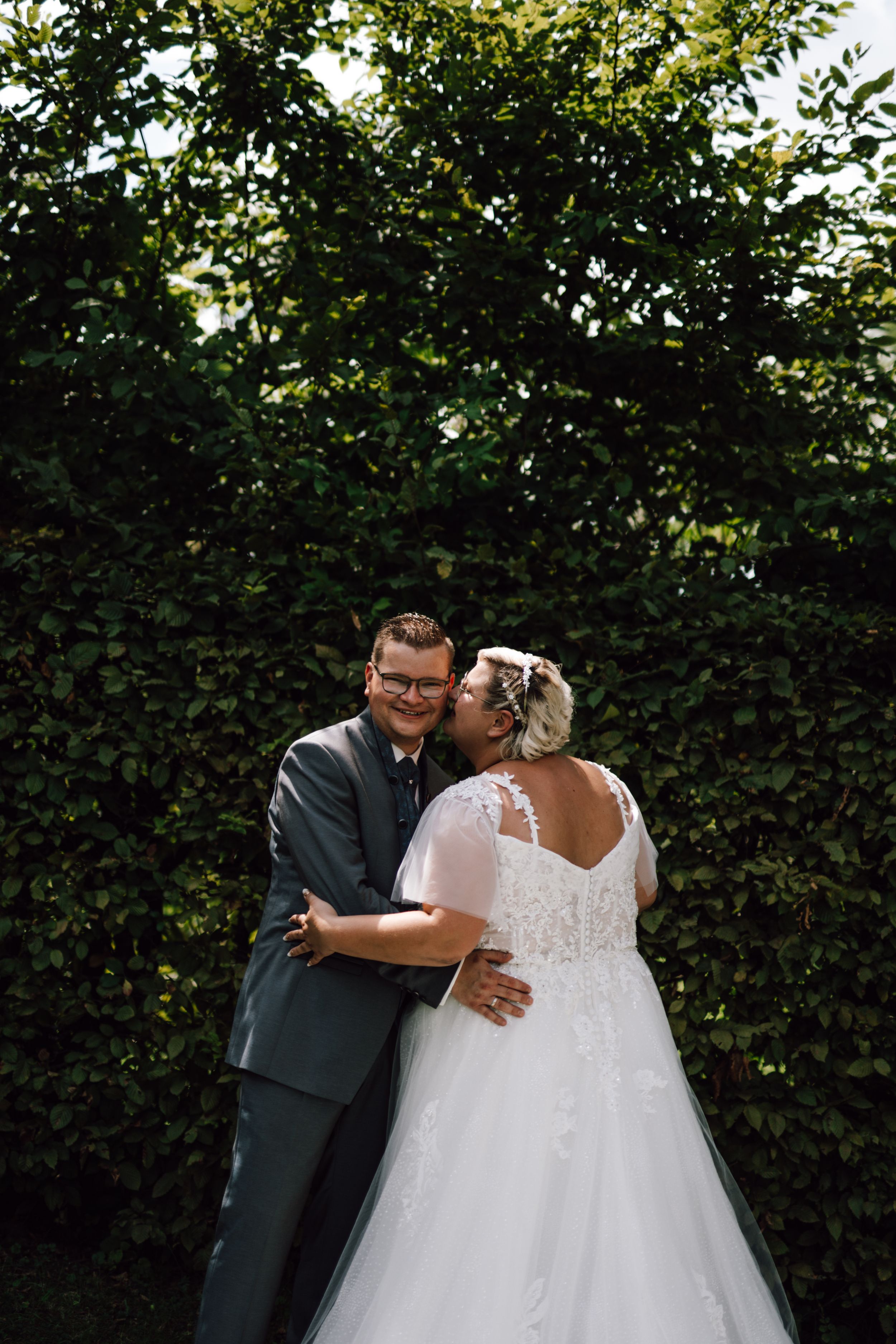 Newlyweds share an intimate moment against a backdrop of green hedges.