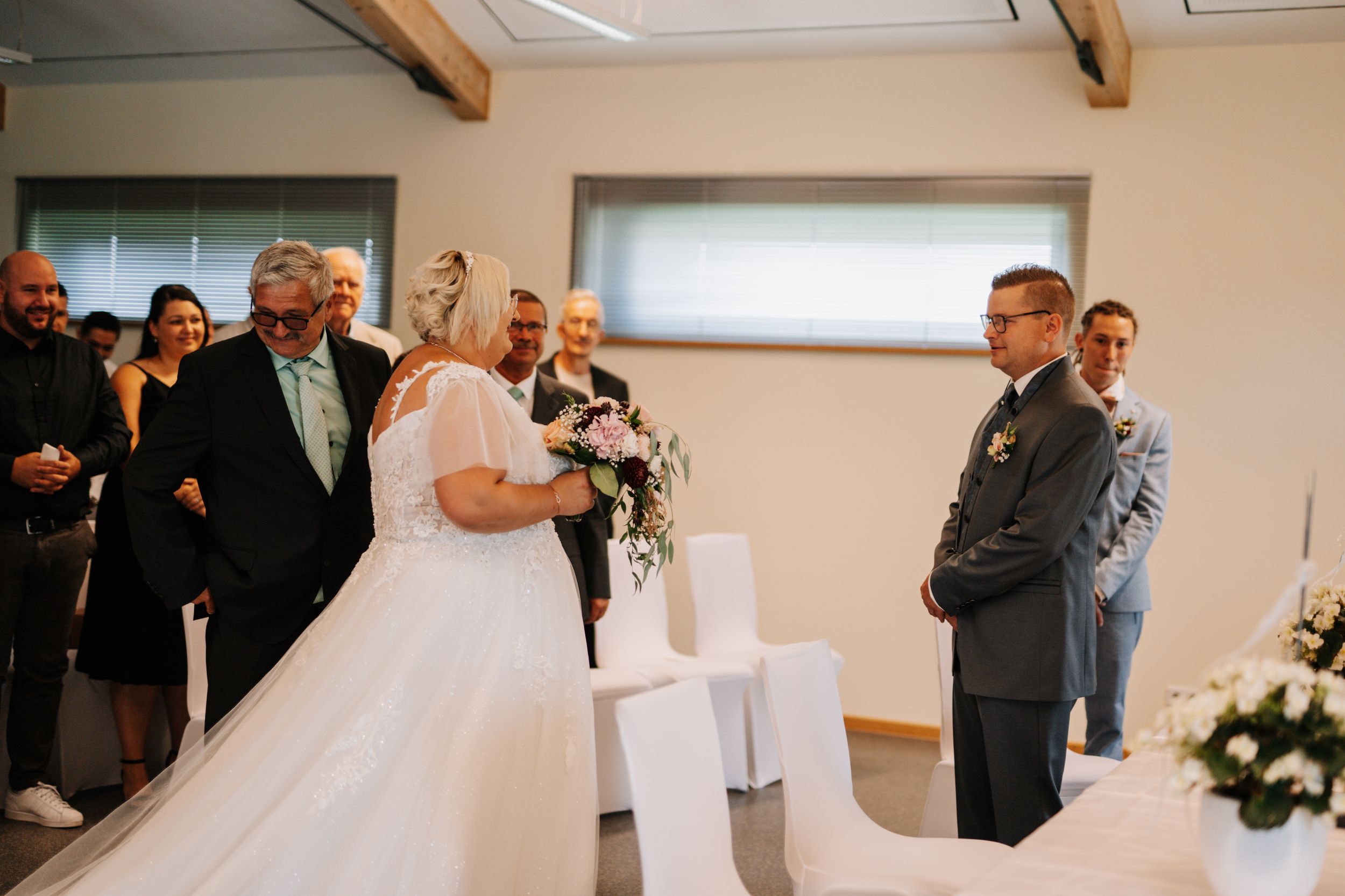 Wedding ceremony in modern indoor venue with white decor and gathered guests.