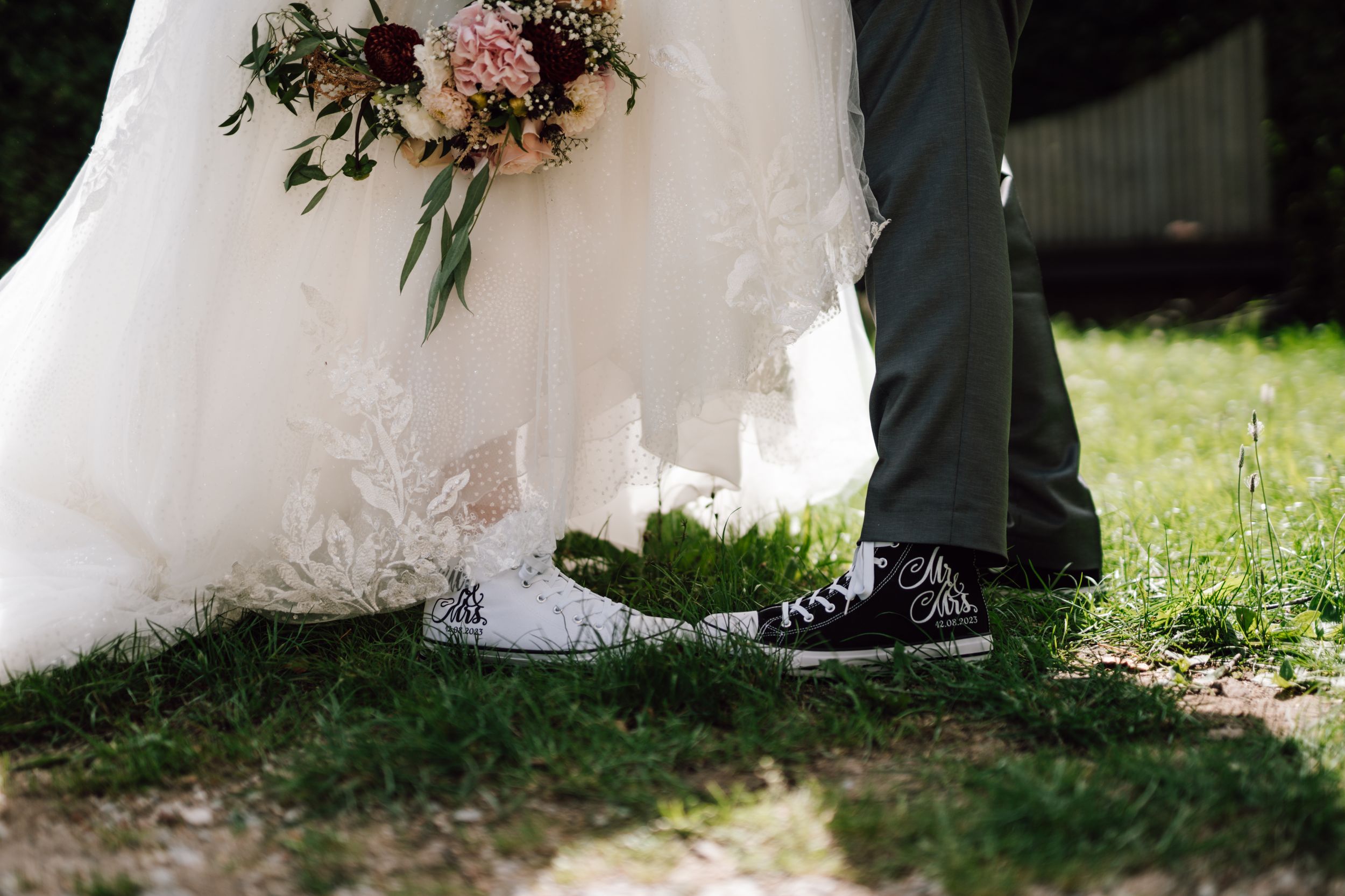 A close-up shot of wedding shoes and dress hem on grass with a pink floral bouquet.