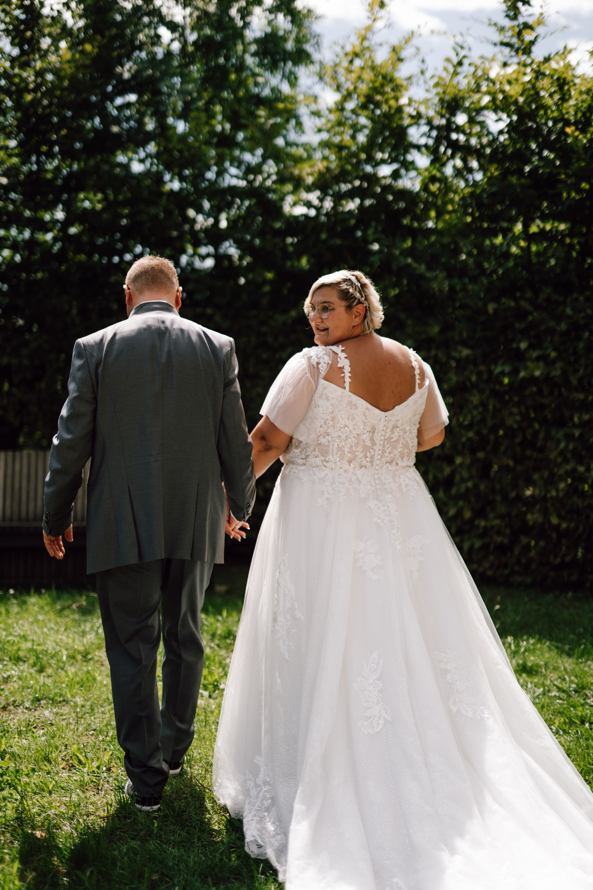 A wedding couple walking hand in hand on grass with the bride wearing a flowing lace gown.