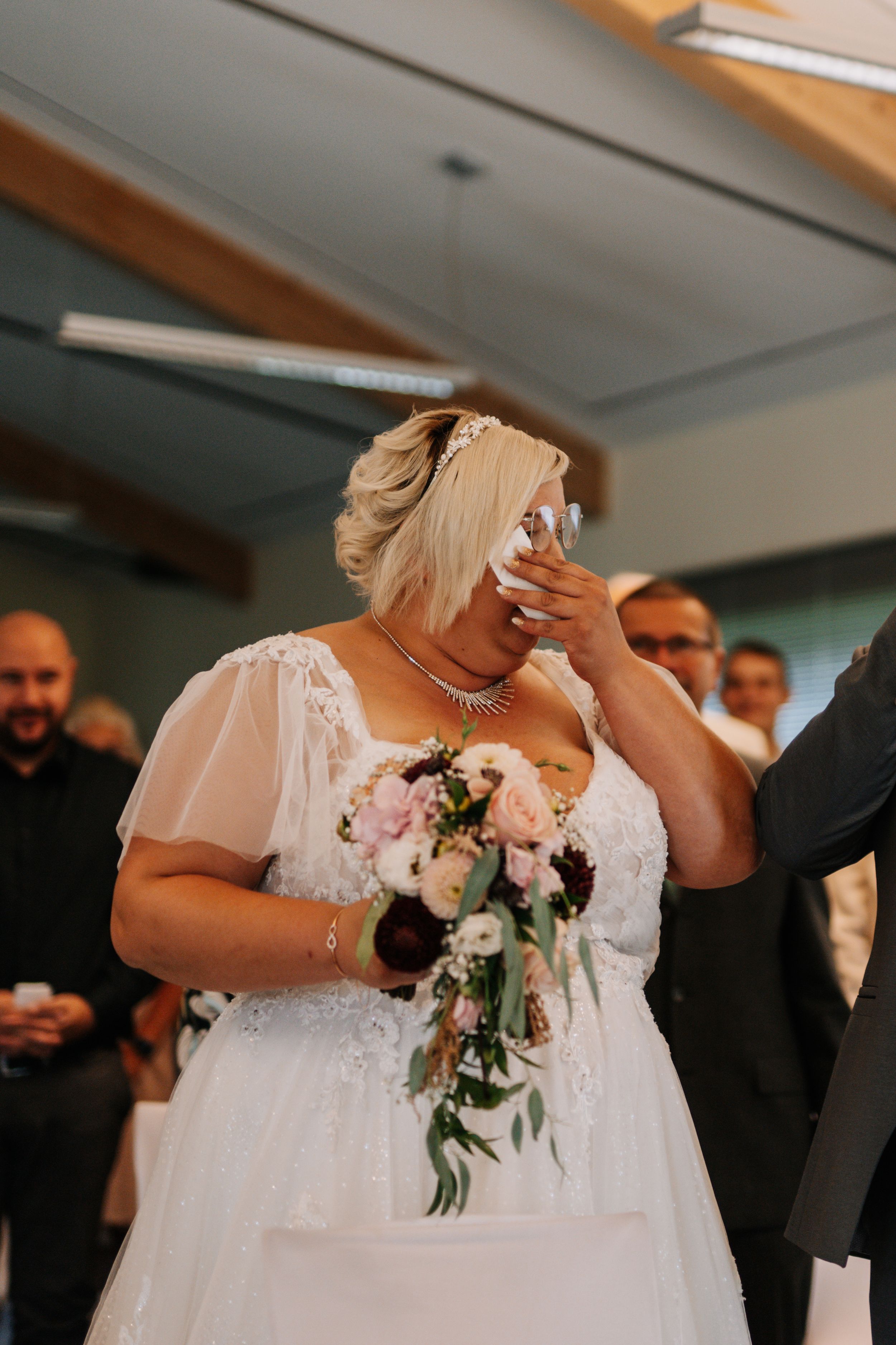 Emotional moment during wedding ceremony as bride in white lace dress holds bouquet of pink and white flowers.
