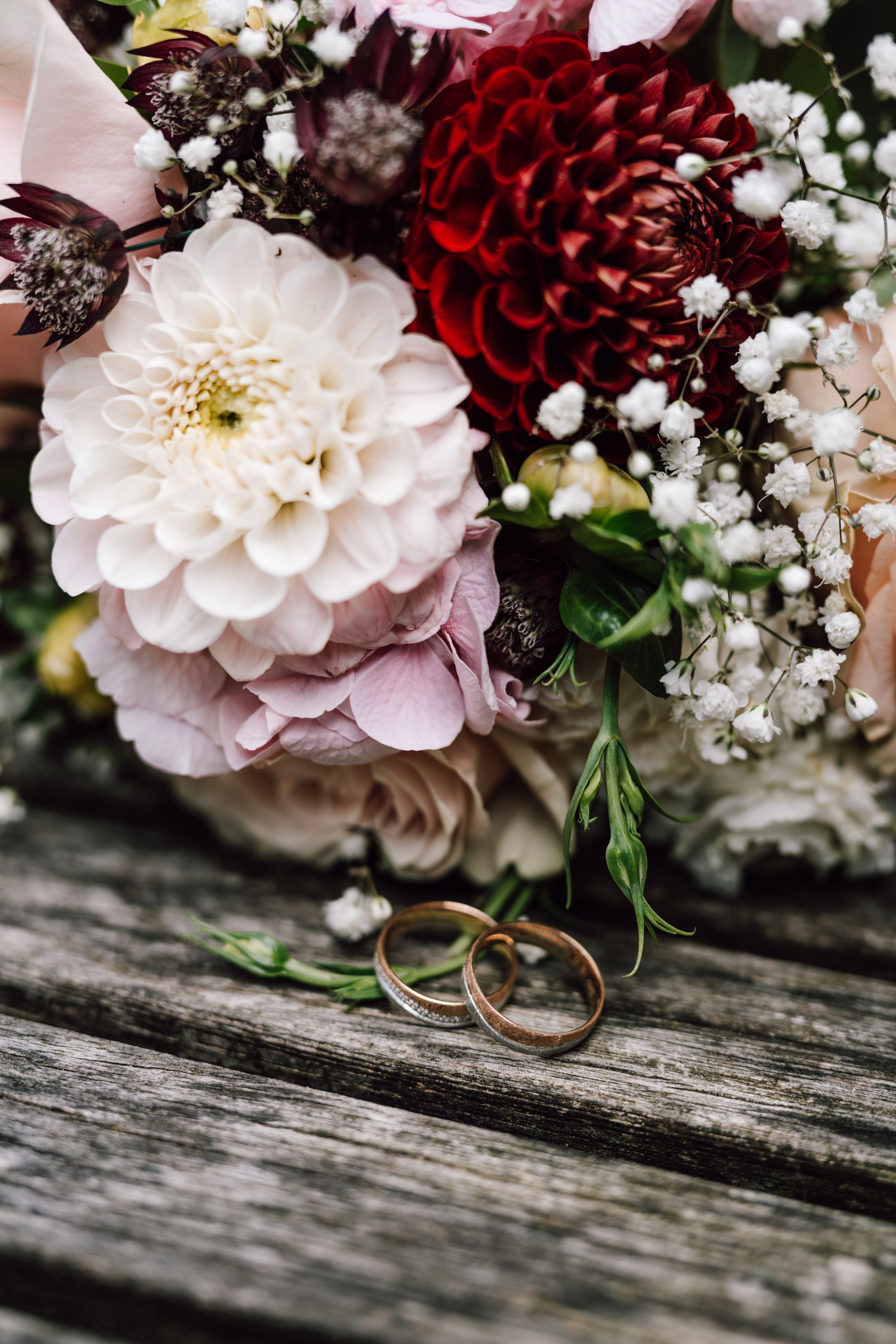Wedding rings beside a stunning bouquet with pink and red dahlias on weathered wood.
