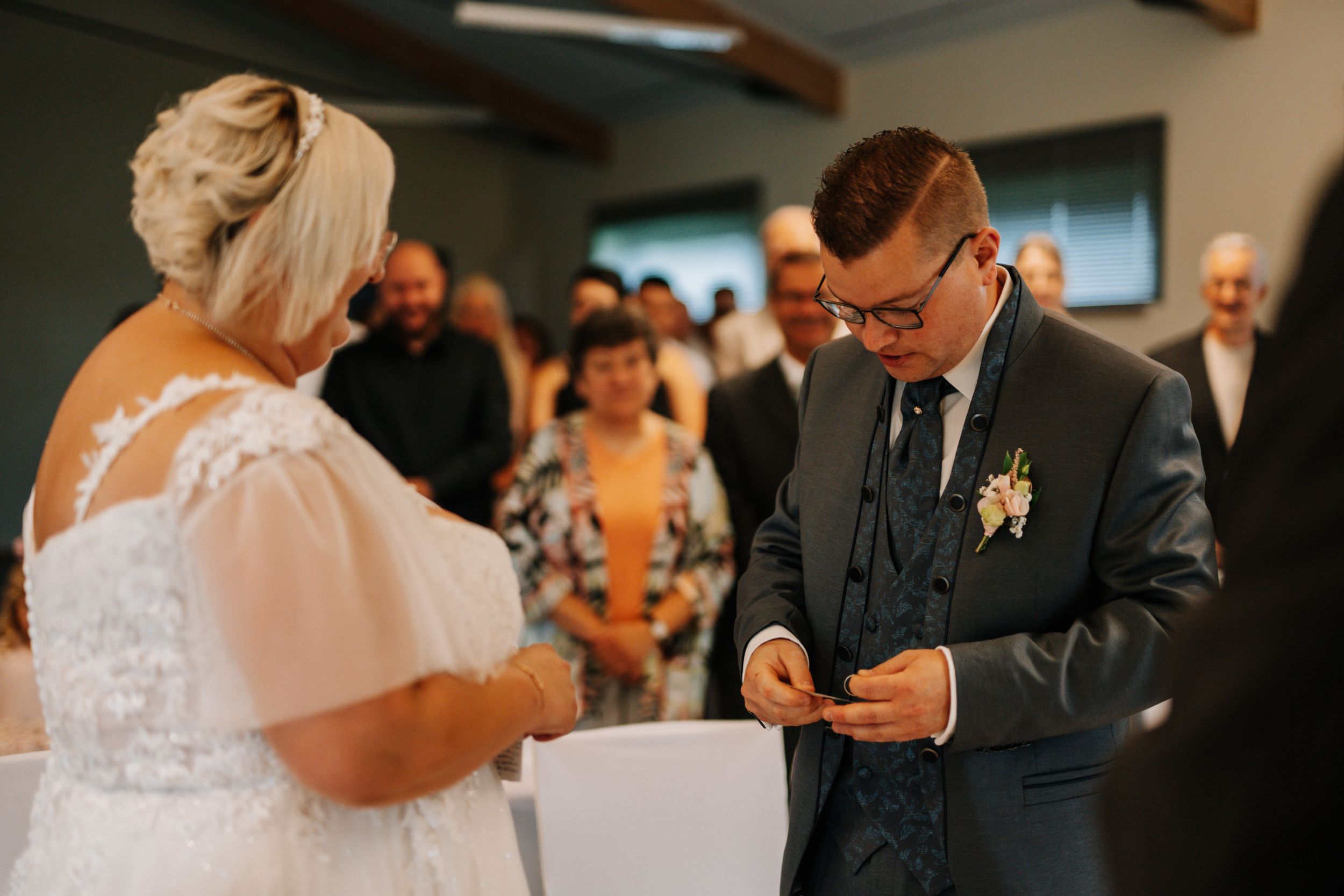 Couple exchanging rings during indoor wedding ceremony with guests watching.