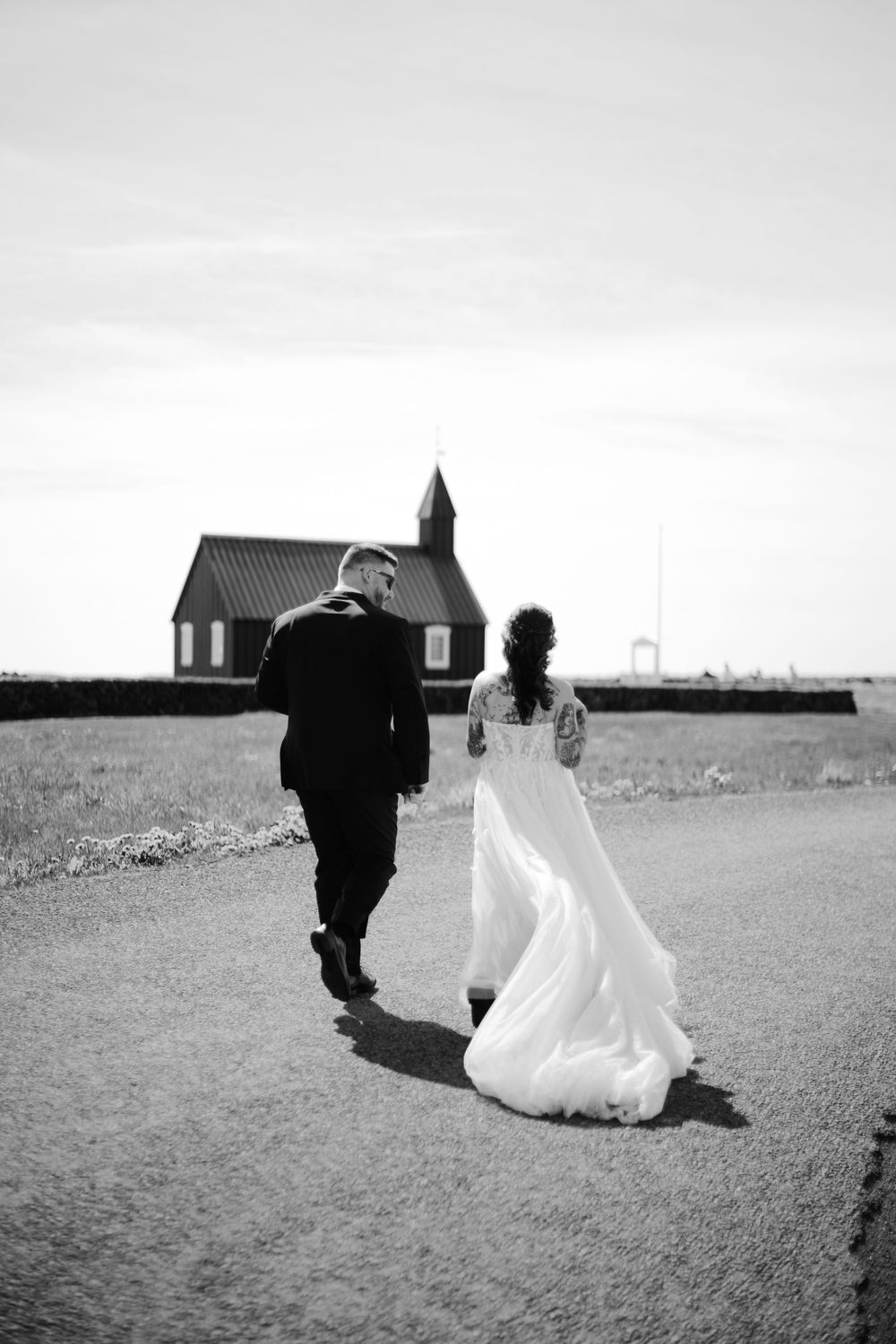 Newlyweds walking together across an open field toward distant buildings.
