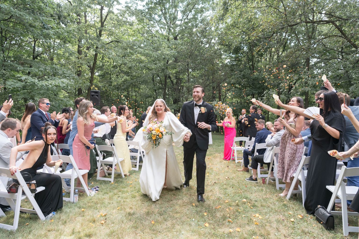 Wedding ceremony guests line pathway with white chairs in wooded outdoor setting during recessional.