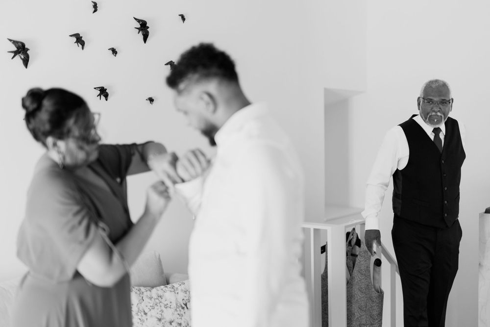 Groom gets ready as family helps him dress in black and white behind-the-scenes wedding preparation photo.