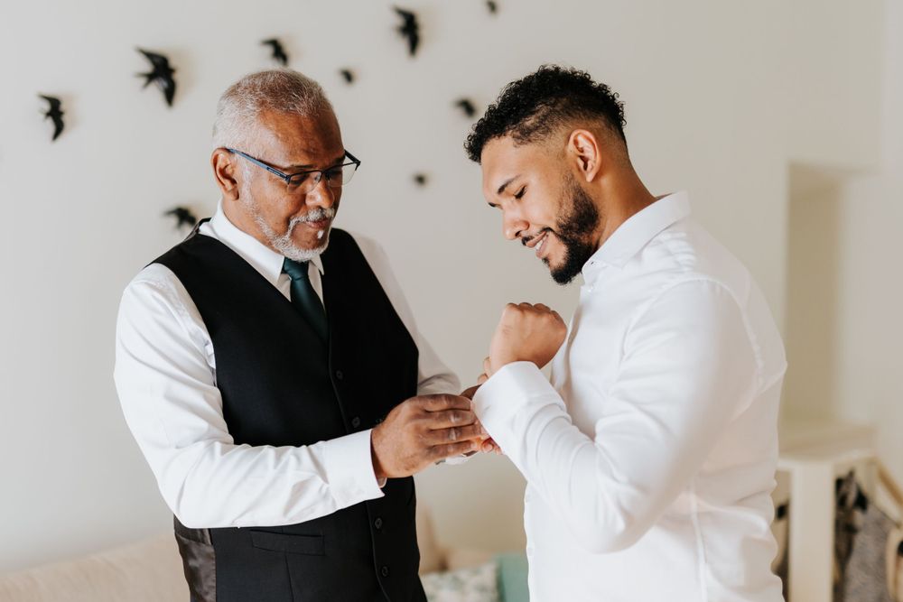 Older man in black vest helping a young groom in white shirt fasten his cufflinks.