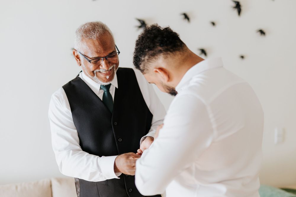 Father smiling while helping his son in a white shirt prepare for his wedding day.