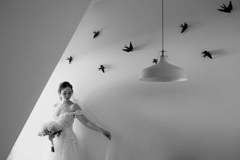 Bride in white gown holds bouquet surrounded by bird wall art in black and white wedding photo.