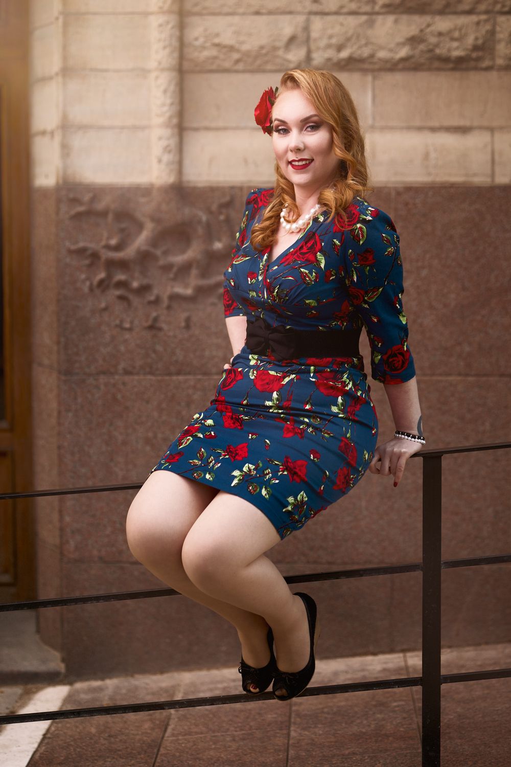 Vintage fashion portrait with floral dress and red hair flower against stone wall background.