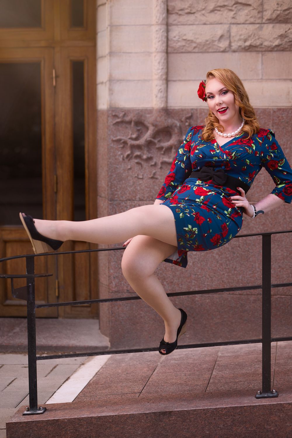 Playful pin-up style pose on railing wearing floral dress and classic heels.