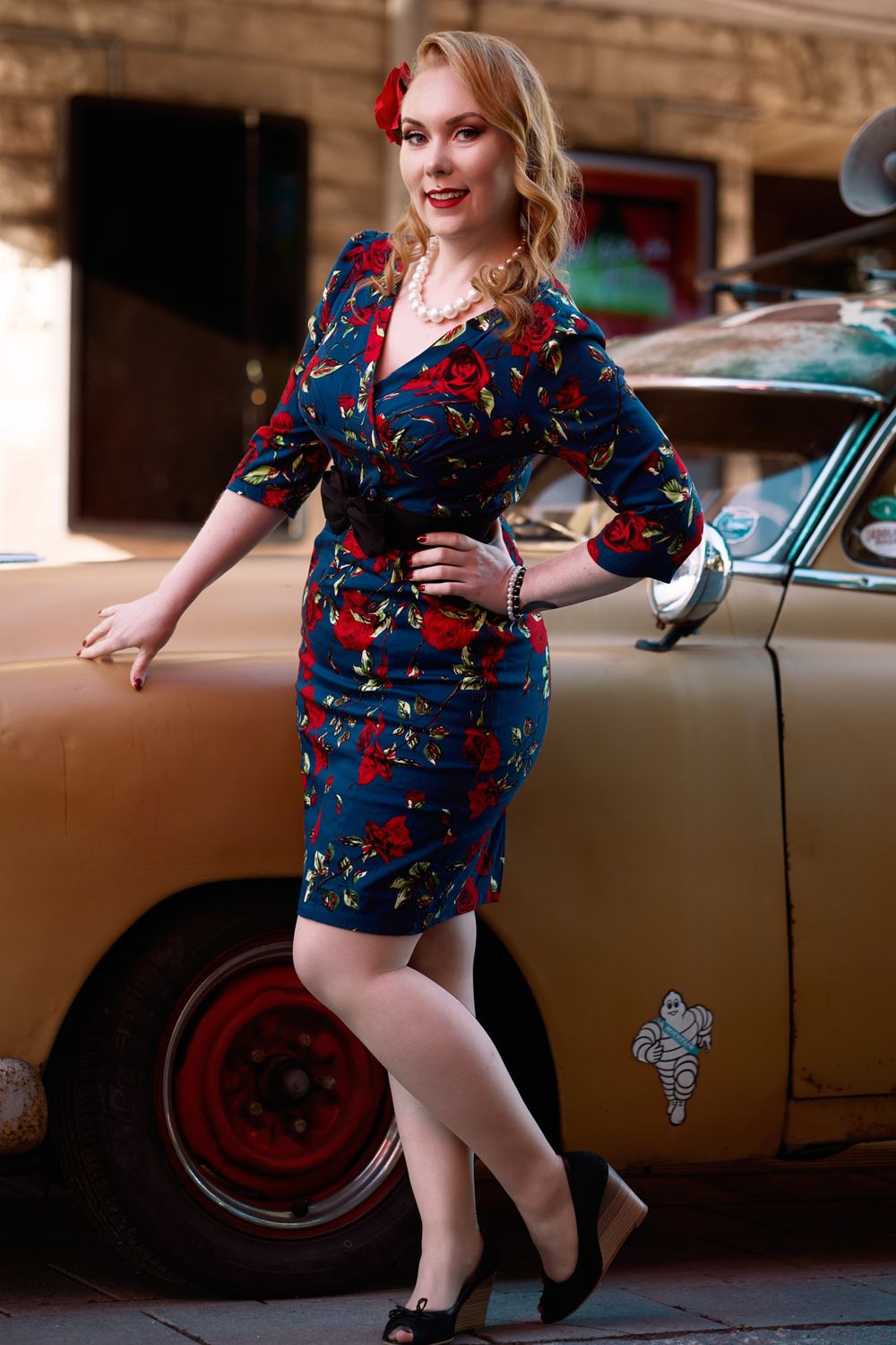Pin-up style fashion pose next to classic brown car featuring floral dress and vintage styling.