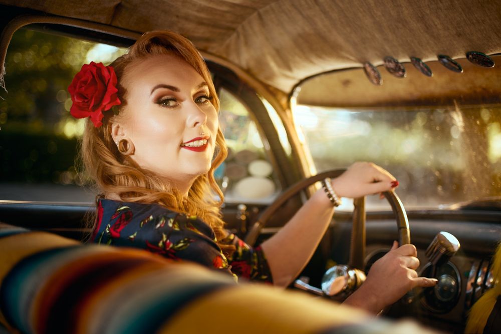 Vintage style portrait inside classic car with red flower hair accessory and retro styling.
