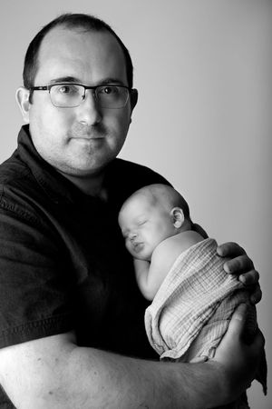 Black and white portrait of a man in glasses tenderly cradling a sleeping swaddled infant.