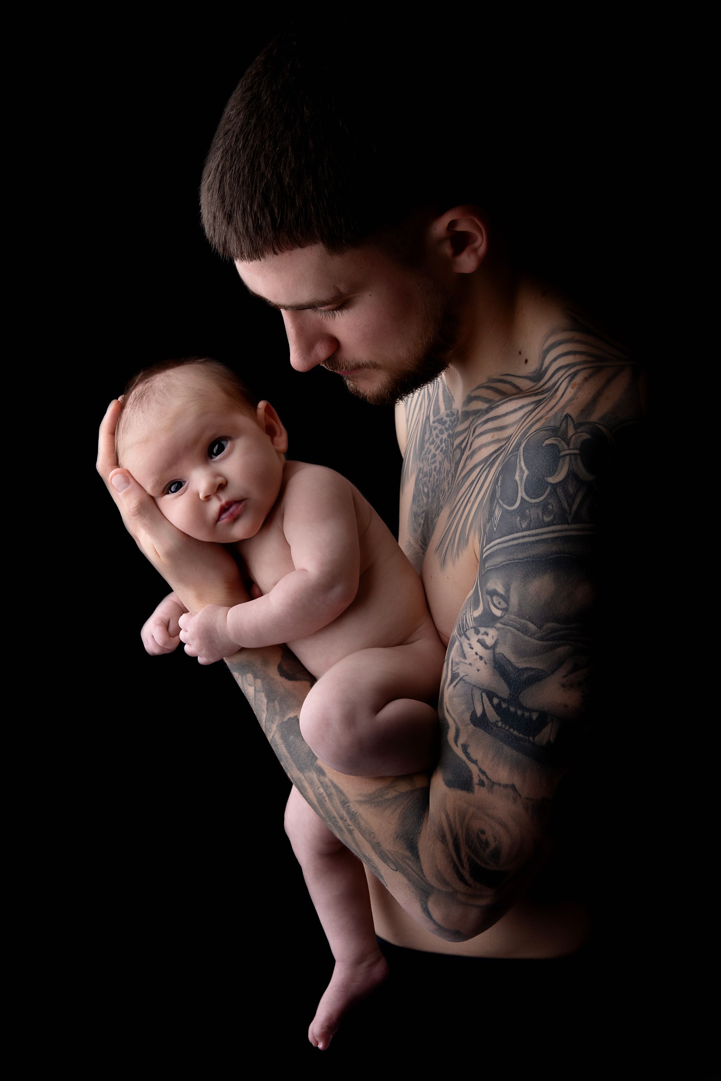 A tender black and white portrait of a tattooed parent cradling and gazing down at a nude baby against dark background.