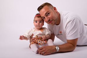 Father and baby in white clothes posing together on light background during studio photoshoot.