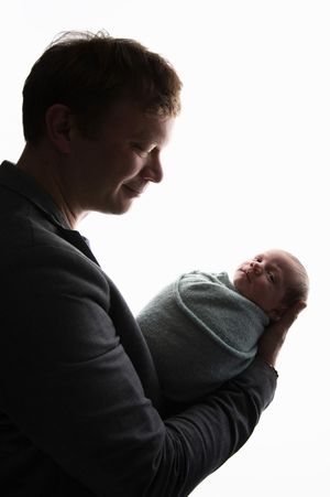 Silhouette photo of adult cradling swaddled newborn baby against bright background.