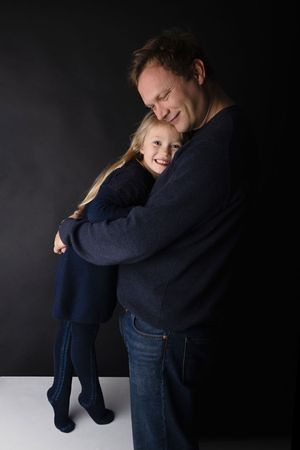 Candid moment of adult embracing young child in navy outfits against dark backdrop.