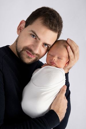 Studio portrait of adult in dark sweater gently holding sleeping swaddled newborn.