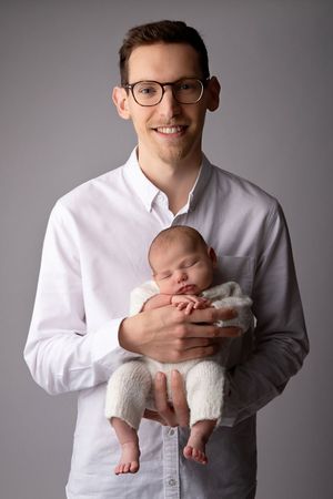 A portrait of a person in glasses and white shirt holding a sleeping newborn baby.