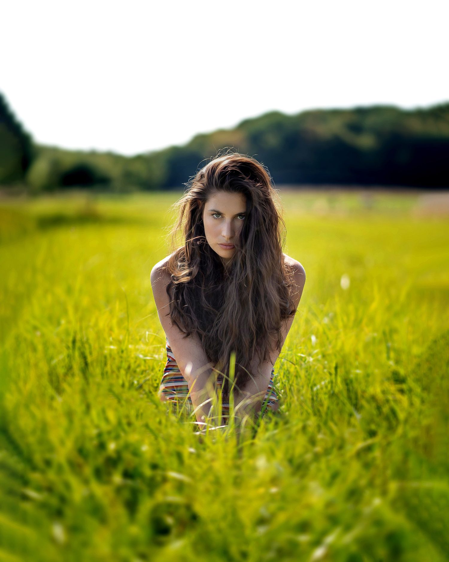Woman with long dark hair kneeling in lush green meadow with forest background.