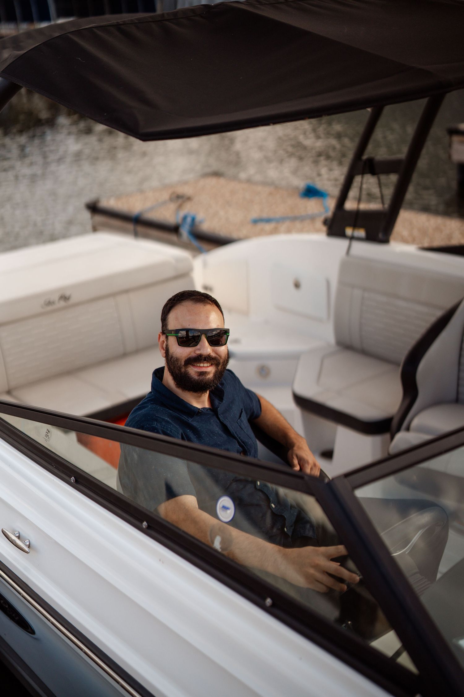 Person wearing sunglasses and a navy shirt sits at the helm of a white boat under a canopy.
