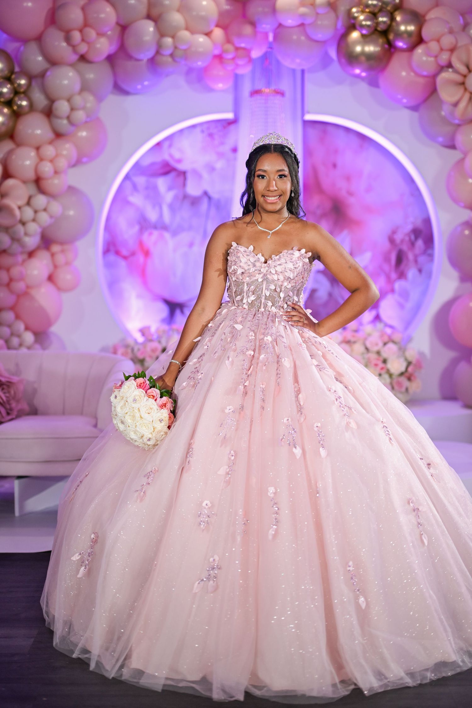 Young woman in a blush pink ball gown with floral appliques holds bouquet at quinceañera celebration.