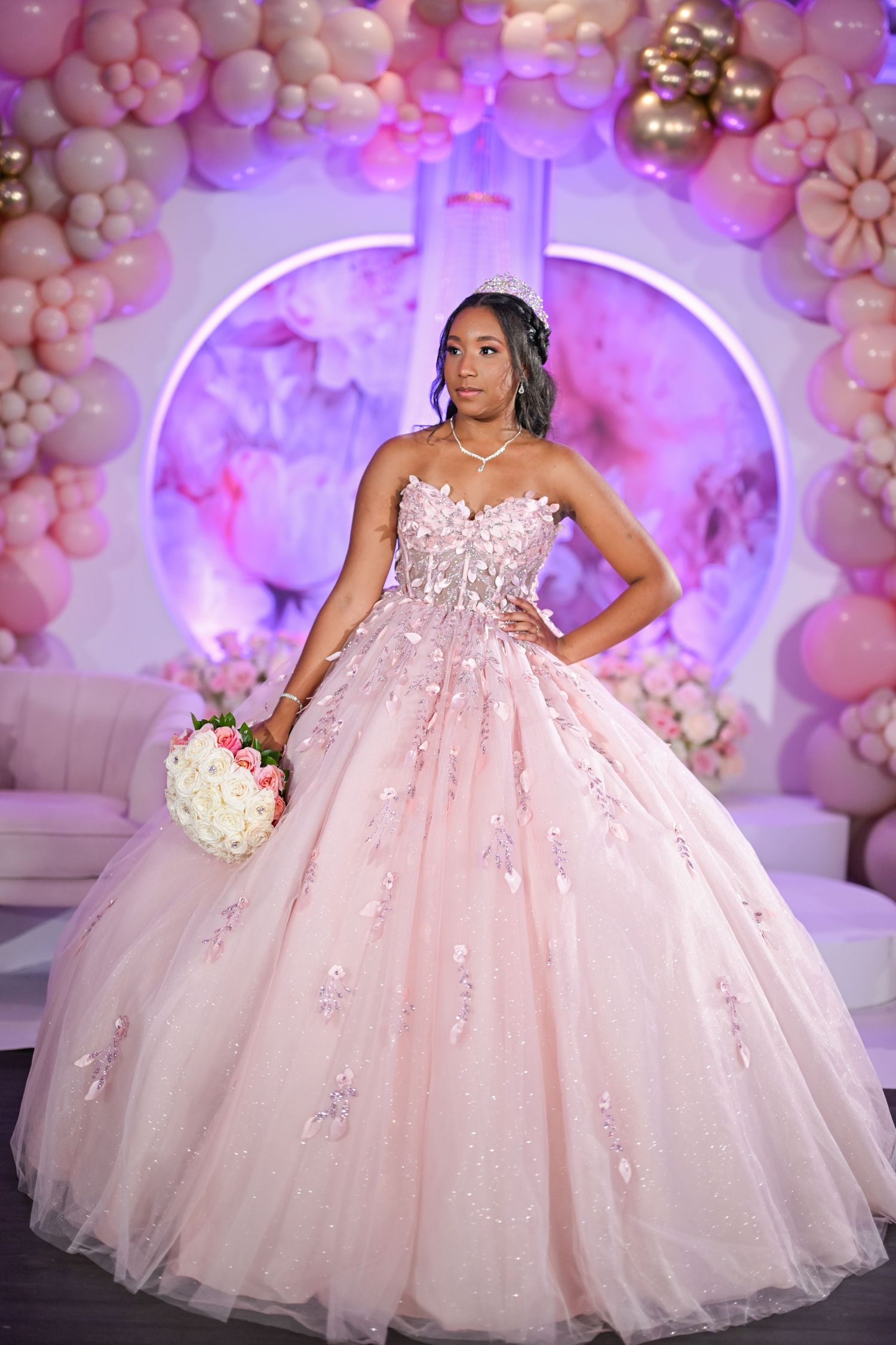 Young woman in a blush pink ball gown with floral embroidery holds bouquet at quinceañera celebration.