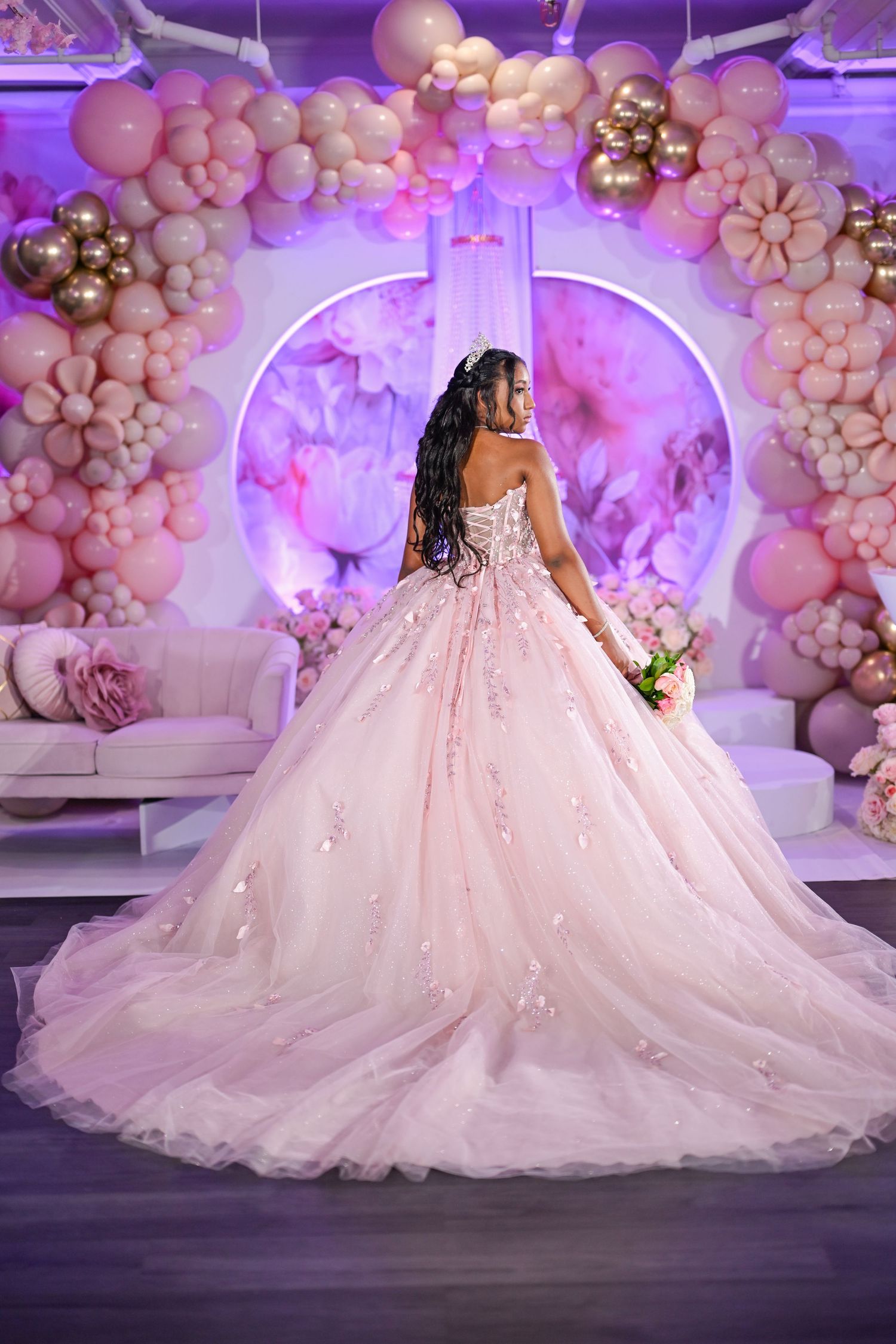 Young woman in a blush pink ball gown quinceañera dress poses before a pink balloon arch decor.